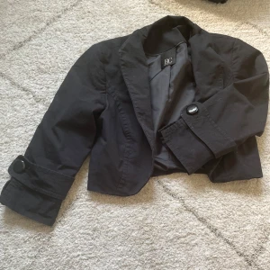 Svart croppad kavaj från Best Connections - Svart  kavaj/ bolero från Best Connections med snygga knappar på ärmsluten och klassisk krage och trekvartärmad. Perfekt för dig som vill ha en stilren och trendig look. Kavajen har en figurnära passform och är fodrad för extra komfort.