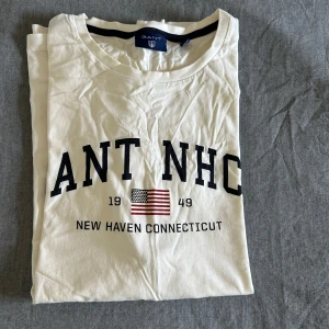 Vit Gant t-shirt med tryck - Vit t-shirt från Gant med svart tryck 'GANT NHCT', amerikansk flagga och texten 'New Haven Connecticut 1949' på bröstet. Klassisk rund hals och korta ärmar. Skön och mjuk bomull, perfekt till jeans eller shorts.