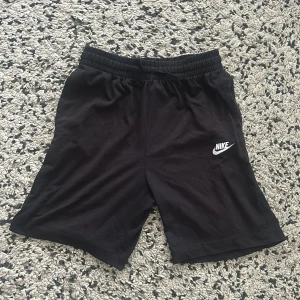 Svarta Nike shorts - Svarta shorts från Nike med klassisk vit logga på ena benet. De har elastisk midja med snörning för skön passform och är gjorda i mjukt bomullsmaterial. Perfekta för träning eller chill dagar. Enkla och stilrena med normal passform.