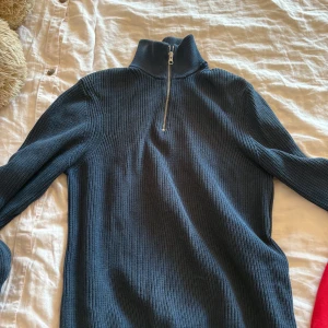 Mörkblå stickad half zip tröja - Säljer en mörkblå stickad tröja med hög krage och half zip-dragkedja. Tröjan har ribbad struktur och långa ärmar, perfekt för lager på lager under kyliga dagar. Enkel och stilren design som funkar till det mesta. Den är även lite stor i storleken
