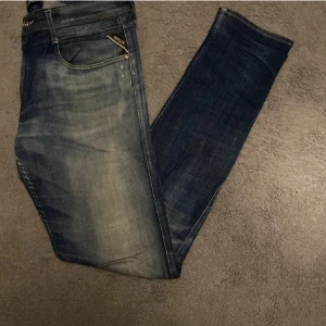 Replay Anbass jeans blå herr - Snygga Replay Anbass jeans i klassisk blå tvätt med slitningar och detaljerade sömmar. Modellen har normal passform och raka ben, med fem fickor och Replay-logga på myntfickan. Perfekta för en avslappnad och trendig look.