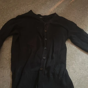 Svart kofta från Lindex XS - En klassisk svart kofta från Lindex i storlek XS. Koftan har långa ärmar, knappar framtill och ribbade muddar vid nederkanten. Perfekt att slänga över en t-shirt eller linne för en chill look.