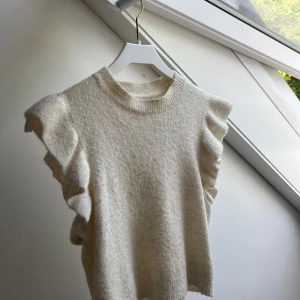 Vit ärmlös stickad tröja Noisy May - Supersöt vit stickad tröja från Noisy May med volangdetaljer på axlarna. Tröjan är ärmlös och har en rund halsringning. Perfekt att styla med jeans eller kjol för en trendig look. Säljer pga att jag har en likadan och den kommer inte till användning 