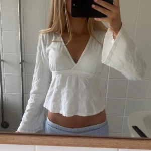 Vit omlottopp med v-ringning - Superfin vit omlottopp med v-ringning och långa, vida ärmar. Toppen har en lätt croppad passform och är sydd i ett tunt, luftigt bomullstyg. Perfekt för vår och sommar när du vill ha en fräsch och stilren look.