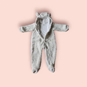 Vit teddyjacka med öron från H&M - Supergullig vit teddyjacka från H&M i storlek 74, med huva och små öron för extra söt vibe. Overallen har dragkedja framtill och är gjord i mjukt fleece-material. Perfekt för kyliga dagar när du vill att din lilla ska vara både varm och stylish.