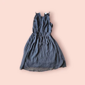 Svart  kortklänning med prickigt mönster - Supersöt mörkgrå kortklänning med tunna axelband och elastisk midja. Klänningen har ett diskret prickigt mönster i silver och är tillverkad i ett lätt, luftigt material som ger en flowy känsla. Perfekt för dig som gillar en enkel men ändå unik stil.