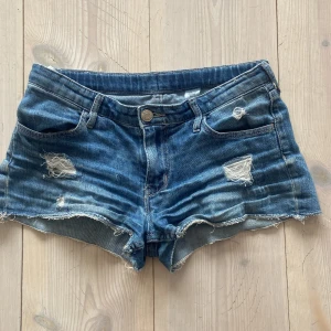 Blåa jeansshorts från &Denim - Snygga blå jeansshorts från &Denim med slitna detaljer och fransig kant. Klassisk femficksmodell med låg midja och dragkedja. Shortsen är i storleken 146 men jag skulle personligen säga att de skulle kunna funka att ha upp till 152. 