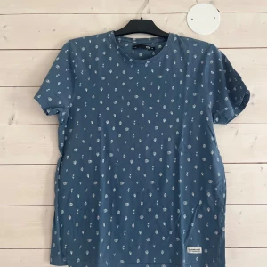 Blå t-shirt med vita blommor från lager 157 - Blå t-shirt från lager 157 med små vita blommor och blad som mönster över hela plagget. Klassisk rund halsringning och korta ärmar. Skön och avslappnad passform, perfekt till jeans eller shorts. Tillverkad i mjuk bomull.