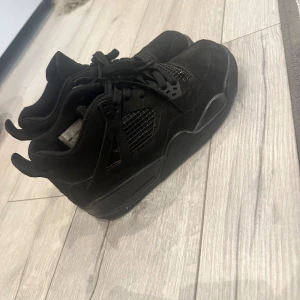 Svarta Nike Air Jordan 4 sneakers - Säljer ett par helsvarta Nike Air Jordan 4 sneakers med klassisk meshpanel och coola detaljer. Skorna har snörning, rund tå och en chunky sula. Ovandelen är i mocka och syntet, vilket ger en stilren och sportig vibe. Perfekta för dig som gillar streetwear och vill sticka ut. Storlek 39