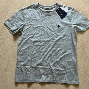 Grå slim fit t-shirt Polo Ralph Lauren - Helt ny! Snygg grå t-shirt från Polo Ralph Lauren i bomull med klassisk rund hals och broderad marinblå logga på bröstet. Modellen är slim fit och har korta ärmar.