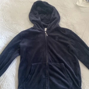 Mörkblå Juicy Couture hoodie i velour - Mysig mörkblå hoodie från Juicy Couture i mjuk velour. Tröjan har dragkedja framtill, huva och känguruficka. Perfekt för chill dagar och har en diskret JC-logga på bröstet. Klassisk och tidlös modell som är superbekväm.