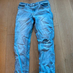 Ljusblå skinny jeans med hål på knät - Säljer ett par ljusblå skinny jeans med klassisk femficksdesign och ett snyggt hål på ena knät. Jeansen har normal midja och är tillverkade i stretchig denim för en tight passform. Perfekta för en avslappnad och trendig look.