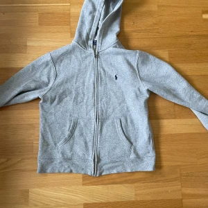 Grå hoodie från Polo Ralph Lauren - Klassisk grå hoodie från Polo Ralph Lauren med dragkedja framtill, huva och två fickor. Tröjan har det ikoniska lilla broderade logotypen på bröstet och är tillverkad i mjuk bomullsmix. Perfekt för en chill och stilren look. Skick 9/10