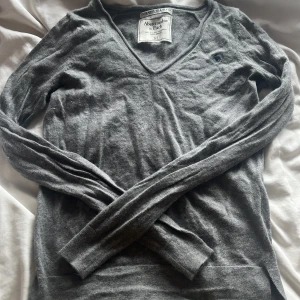 Grå v-ringad tröja Abercrombie XS - Mjuk och tunn grå tröja från Abercrombie & Fitch i kashmir- och silkesblandning. V-ringad modell med lång ärm och liten broderad logga på bröstet. Perfekt för lager-på-lager och chill vibes.