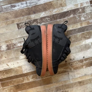 Svarta DC sneakers  - STRL 43 Snygga svarta sneakers från DC Shoes med kraftig brun sula och grova svarta skosnören. Skorna har DC-logga i mocka på sidan och en stjärndetalj. Perfekta för dig som gillar streetstyle och skatekänsla.