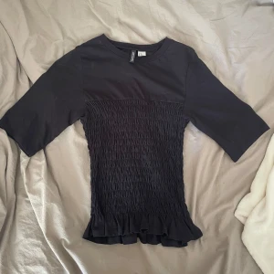 Svart smockad topp från hm - Supersnygg svart topp från hm med smockad midja och volangkant nertill. Toppen har rund halsringning och halvlånga ärmar. Perfekt för dig som gillar en figurnära passform med coola detaljer.