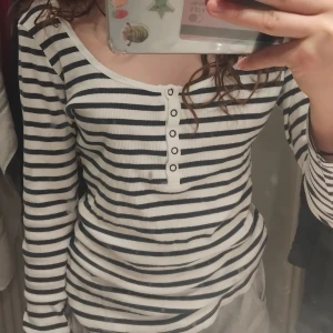 Randig långärmad tröja med knappar - Säljer en ribbad långärmad tröja med marinblå och vita ränder. Tröjan har en snygg v-ringning med dekorativa knappar framtill. Perfekt för dig som gillar en klassisk men chill stil.