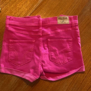 Rosa shorts från Hollister W27 - Snygga rosa shorts från Hollister med klassisk femficksdesign och uppvikta benslut. Modellen har normal passform och är tillverkad i mjuk bomull, perfekta för varma sommardagar. Tydlig Hollister-logga bak på linningen.