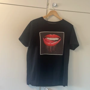 Svart Limitato t-shirt med läppar - Svart t-shirt från Limitato med ett stort tryck av röda läppar och tänder på bröstet. T-shirten har klassisk passform, rund hals och är kortärmad. Materialet är mjukt och skönt, perfekt för dig som vill sticka ut med en cool och edgy look.