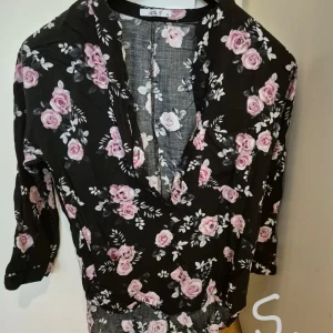 Svart blommig skjorta med rosa rosor - Supersöt svart skjorta från Hollister med rosa och vita rosor över hela tyget. Skjortan har v-ringning, knappar framtill och trekvartsärm. Perfekt för dig som gillar romantiska och feminina vibes.