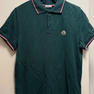 Grön Moncler pikétröja med krage - Snygg mörkgrön pikétröja från Moncler med klassisk krage och knäppning framtill. Tricolore-detaljer på krage och ärmslut samt broderad Moncler-logga på bröstet. Perfekt för en stilren och sportig look.