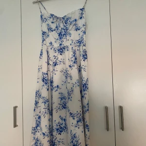 Vit och blå blommig klänning Gina Tricot - Superfin vit klänning med blått blommönster från Gina Tricot. Klänningen har smala axelband, snörning i ryggen och en söt knytdetalj framtill. Den är midilång med en flowy kjol och markerad midja, perfekt för sommarens alla tillfällen. Lappen sitter kvar, helt oanvänd, Endast testad☺️