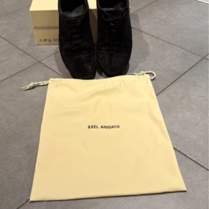 Svarta sneakers från Axel Arigato - Stilrena svarta sneakers från Axel Arigato i mocka med platt sula och rund tå. Skorna har klassisk snörning och en minimalistisk design som passar till det mesta. Levereras med originalkartong och dustbag.