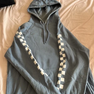 Blå hoodie från Vans med rutiga detaljer - Säljer en blå hoodie från Vans med vita rutiga ränder längs ärmarna och broderad Vans-logga på bröstet. Tröjan har huva med snörning och är tillverkad i mjuk bomull. Perfekt för dig som gillar streetwear och vill ha en chill stil.