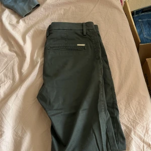 Mörkgröna kostymbyxor från Oscar Jacobson - Snygga mörkgröna kostymbyxor från Oscar Jacobson med klassisk passform. Byxorna har hällor för bälte, diskreta bakfickor och en stilren metallogo ovanför ena fickan. Perfekta för en clean och dressad look.