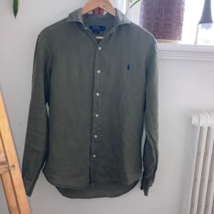 Olivgrön linneskjorta Polo Ralph Lauren - Snygg olivgrön skjorta från Polo Ralph Lauren i linne med klassisk krage och blå broderad logga på bröstet. Slim fit-modell med långa ärmar och knäppning framtill. Perfekt för dig som gillar stilrena och bekväma plagg.