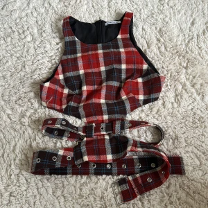 Rutig korsettopp från Rave Review - Säljer en röd, brun och vit rutig korsettopp från Rave Review. Toppen har breda axelband, dragkedja i ryggen och coola bältesdetaljer som korsas bak. Materialet ullblandning. Perfekt för dig som vill sticka ut med edgy vibe.