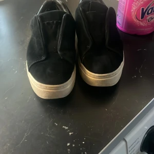 Svarta sneakers med vit sula - Snygga svarta sneakers med en vit sula. Skorna har en stilren design utan snörning och är perfekta för en avslappnad look. De är tillverkade i ett material som liknar mocka och har en bekväm passform. Återsäljer då de inte passade mig