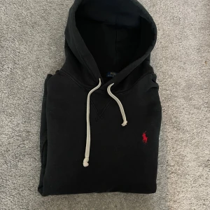 Svart hoodie från Polo Ralph Lauren - Svart hoodie från Polo Ralph Lauren med klassisk röd logga broderad på bröstet. Tröjan har huva med vita snören och är tillverkad i mjuk bomull. Perfekt för en chill och stilren look. Är i storlek L sitter mellan L och M