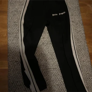 Palm angels byxor  - Svarta mjukisbyxor från Palm Angels med vita sidoränder och logga på höften. Byxorna har dragkedja vid benslut och är i en slim passform. Perfekta för en sportig och trendig look.