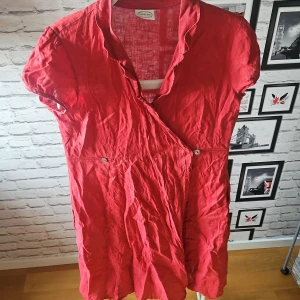 Röd omlottklänning från Newhouse - Snygg röd omlotttklänning från Newhouse i linne med omlott-design och korta ärmar. Klänningen har v-ringning, knappar i midjan och en luftig känsla – perfekt för varma dagar. Samt två fickor på sidorna.