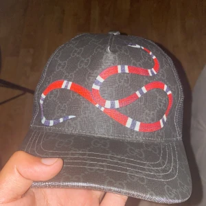 Gucci keps med orm och GG-mönster - Snygg svart Gucci keps med klassiskt GG-monogram och stor röd orm på framsidan. Baksidan har mesh och justerbart band i blått och rött. Kepsen är tillverkad i polyester och har exklusiva detaljer som sticker ut.