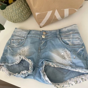 Ljusblå jeansshorts från G-Star - Snygga ljusblå jeansshorts från G-Star med slitna detaljer, nitar och pärlor framtill. Shortsen har fransiga kanter med blommigt tyg på insidan och dubbla knappar i midjan. Perfekta för sommaren och ger en cool, avslappnad vibe.