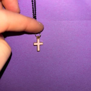 Svart halsband med korshänge - Stilrent svart halsband med ett litet korshänge i guld och vitt. Kedjan är tunn och enkel, medan korset har en klassisk form med guldfärgad kant och vit fyllning. Perfekt accessoar för dig som gillar minimalistiska smycken med en touch av symbolik.