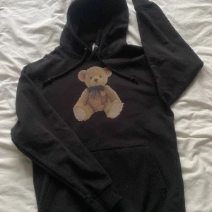 Svart hoodie med nalle från Just Hoods - Svart hoodie från Just Hoods i storlek S med en stor, söt nallebjörn-tryck på bröstet. Klassisk huva med snörning och känguruficka framtill. Perfekt för dig som gillar streetstyle och vill ha något unikt och mysigt i garderoben. nästan aldrig använd, pris kan diskuteras. 