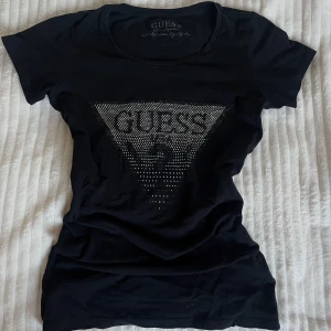 Svart t-shirt från Guess - Svart t-shirt från Guess med ikonisk logga framtill med rhinestones. Tröjan har tight passform och gjord av 95% bomull. Tröjan är ikonisk och ger verkligen y2k vibe💗