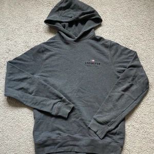 Mörkgrå hoodie från Lexington - Mörkgrå hoodie från Lexington med broderad logga och liten flaggdetalj på bröstet. Klassisk modell med huva och långa ärmar, ribbade muddar och mjukt material som känns skönt mot huden. Perfekt för en chill och avslappnad stil.