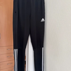 Svarta Adidas mjukisbyxor med vita ränder - Svarta mjukisbyxor från Adidas med klassiska vita ränder längs benen och Adidas-logga på låret. Byxorna har elastisk midja och är gjorda i polyester, perfekta för träning eller chill. Snygg och sportig look med smal passform. 