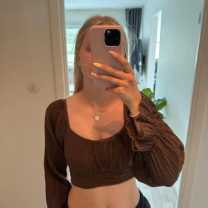 Brun croppad omlottopp med knyt - Supersnygg brun croppad topp från H&M med långa puffiga ärmar och omlottdesign. Toppen har en bred fyrkantig ringning och knyts i ryggen för en extra cool detalj. Tillverkad i ett lätt, krinklat bomullstyg som känns luftigt.