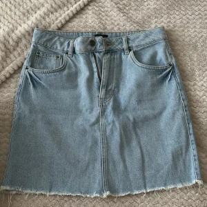 Ljusblå jeanskjol från NLY Jeans - Säljer en ljusblå jeanskjol från NLY Jeans med klassisk femficksdesign, bälteshällor och rå, fransig kant nedtill. Kjolen är kort och har dragkedja och knapp framtill. Perfekt för dig som gillar en avslappnad och trendig stil.