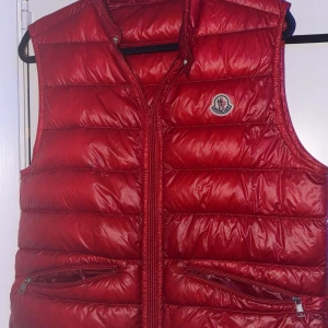 Röd dunväst från Moncler - Snygg röd dunväst från Moncler med klassisk quiltad design och dragkedja framtill. Västen har två praktiska fickor med dragkedja och ikonisk Moncler-logga på bröstet. Perfekt för lager-på-lager och ger en sportig vibe till din outfit.