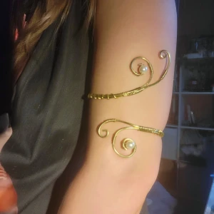 Handgjord Guldfärgat armring med pärldetaljer - Unik armring i guldfärgad metall med två spiralformade ändar, dekorerade med vita pärlor. Snyggt handgjord design som ger en bohemisk och elegant känsla. Perfekt accessoar för att lyfta din outfit.