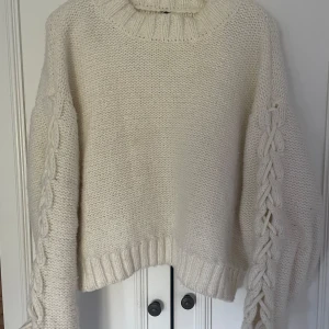 Vit stickad tröja med flätade detaljer - Säljer en mysig vit stickad tröja med grovstickad struktur och flätade detaljer längs ärmarna. Tröjan har en rund halsringning och ribbade muddar vid ärmslut och nederkant. Perfekt för kyliga dagar!