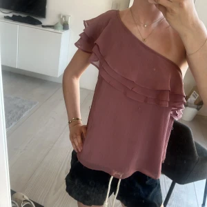 Rosa one shoulder topp med volang - Säljer en söt rosa one shoulder topp med volangdetaljer. Toppen har en luftig passform och är perfekt för varma dagar. Materialet känns lätt och bekvämt mot huden. Passar dig som vill ha en trendig och feminin look.