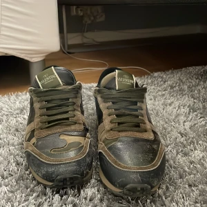 Valentino Rockrunner - Säljer ett par Valentino Rockrunner sneakers med camouflage-mönster i grönt, beige och svart. Skorna har röda detaljer på hälen, snörning och platt sula. Ikoniska nitar på baksidan av sulan och klassisk rund tå.Blev lite suddiga bilder sorry.