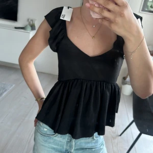 Svart peplumtop med v-ringning - Svart topp med v-ringad hals och peplumskärning. Toppen har korta ärmar och är tillverkad i ett lätt, något strukturerat material. Perfekt att styla med jeans eller kjol.helt ny med prislapp 